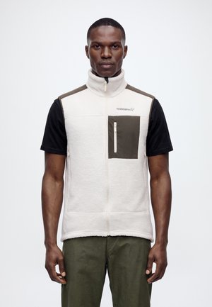 Un hombre luce un chaleco polar blanco con cremallera y un bolsillo oscuro en el pecho, sobre una camiseta negra y unos pantalones verde oliva, con un fondo liso.