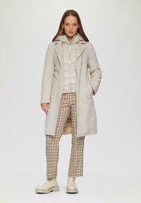 Beige wollen overjas over een lichte pufferjack; geruite broek in beige, bruin en groen; crèmewitte enkellaarsjes; gladde texturen.