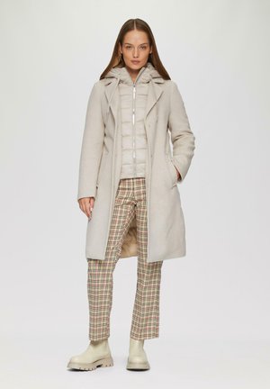 Manteau en laine beige par-dessus une veste rembourrée légère ; pantalon à carreaux beiges, marron et verts ; bottines crème à la cheville ; textures lisses.