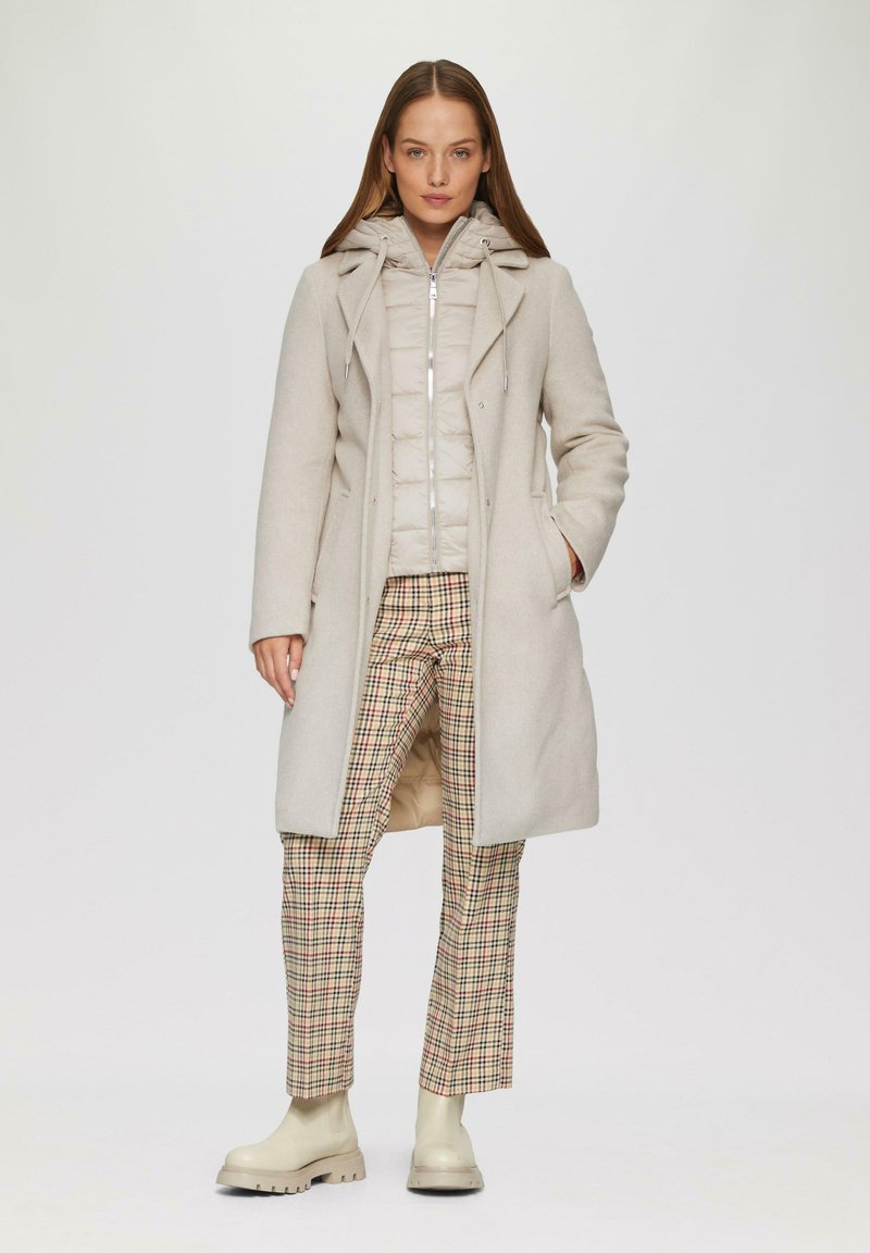 Beige wollen overjas over een lichte pufferjack; geruite broek in beige, bruin en groen; crèmewitte enkellaarsjes; gladde texturen.