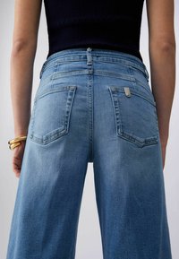 Naine, kes kannab kõrge vöökohaga, laia säärega siniseid teksapükse ja tumedat varrukateta topi, vasakul randmel kuldseid käevõrusid.