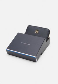 Schwarzes Lederportemonnaie mit goldenem Logo, teilweise sichtbar in einer blauen Box mit einem weißen und roten Streifen. Glatte Textur, Reißverschluss.