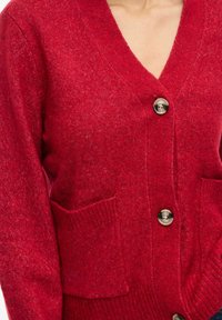Crvena vunena kardigan s V-izrezom, dva bež gumba zakopčana, i prednji džepovi, nosi ga osoba.