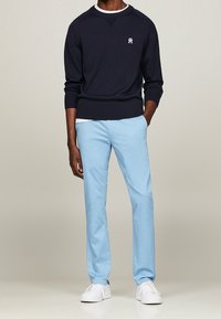 Granatowy sweter z dzianiny z prążkowanymi mankietami i dołem, z małym logo. Jasnoniebieskie spodnie, gładka tekstura. Białe sneakersy z sznurowadłami.