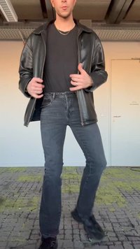 Bărbat purtând o jachetă de piele neagră, tricou negru, blugi închiși la culoare și pantofi negri, stând în interior pe un podea cu model.