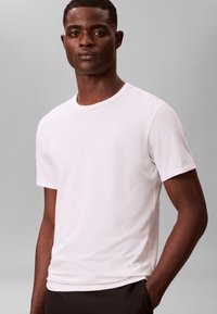 Λευκό βαμβακερό T-shirt με κοντά μανίκια, στρογγυλή λαιμόκοψη και λεία υφή, συνδυασμένο με σκουρόχρωμο παντελόνι. Απλός σχεδιασμός, χωρίς ορατά μοτίβα.