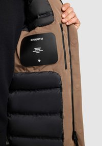 Brun jacka med svart vadderat inre. Har en slät tygsida, dragkedja och en tryckt svart etikett med märkesdetaljer.