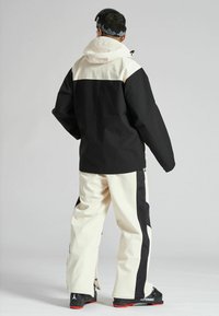 Chaqueta de esquí negra y crema con capucha, que presenta un diseño de color dividido. Combinada con pantalones de esquí crema y negros. Textura suave, estilo funcional.