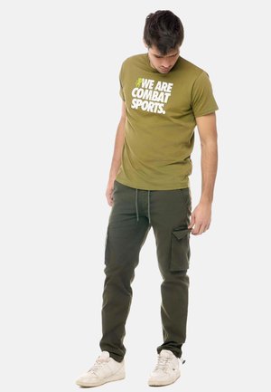 Uomo che indossa una t-shirt verde oliva “We Are Combat Sports”, pantaloni cargo verde scuro e sneakers bianche, con lo sguardo rivolto verso il basso. Sfondo semplice.