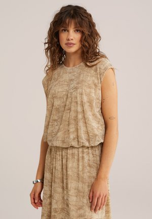 Mujer con cabello rizado y castaño, vestida con una blusa sin mangas beige con textura y una falda a juego, sobre un fondo liso.