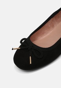 Ballerine noire à bout arrondi, nœud tressé noir décoratif avec embouts dorés et doublure intérieure beige douce.
