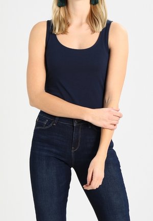 Kobieta w ciemnoniebieskim topie bez rękawów i ciemnoniebieskich dżinsach, stojąca z założonymi rękami na jasnym, jednolitym tle.