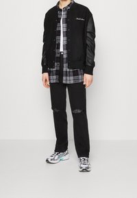 Černá bomber bunda se semišovým tělem a koženými rukávy, vrstvená přes černou a bílou kostkovanou košili, v kombinaci s potrhanými černými džíny a stříbrnými teniskami.