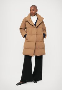 Nainen, jolla on ajeltu pää, käyttää vaaleanruskeaa toppatakkia valkoisen paidan päällä, mustia leveitä housuja ja mustia matalia kenkiä, seisten kädet taskuissa.