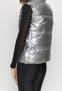 Silverfärgad vadderad väst med glänsande, reflekterande material, ärmlös design och stygnsektioner. Buren över en svart långärmad topp och leggings.