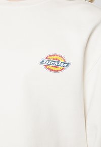 Vêtement blanc avec un petit logo Dickies usé en rouge, jaune et bleu sur la partie supérieure de la poitrine.