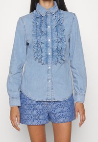 Ljusblå denimskjorta med volangdetalj fram, spetsig krage, knäppning med knappar och långa ärmar. Bärs med blå mönstrade shorts.