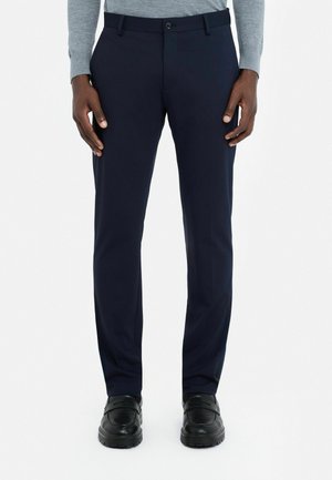 Marinebroeken van gladde stof, met een rechte pasvorm, riemlussen en een enkele knoopsluiting, gedragen met zwarte schoenen.