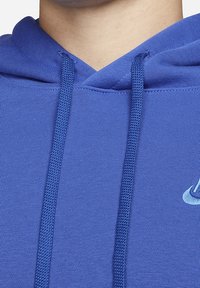 Felpa blu con un tessuto testurizzato, dotata di un cappuccio con cordino e un piccolo logo sul petto. Vista ravvicinata del colletto e delle cuciture.