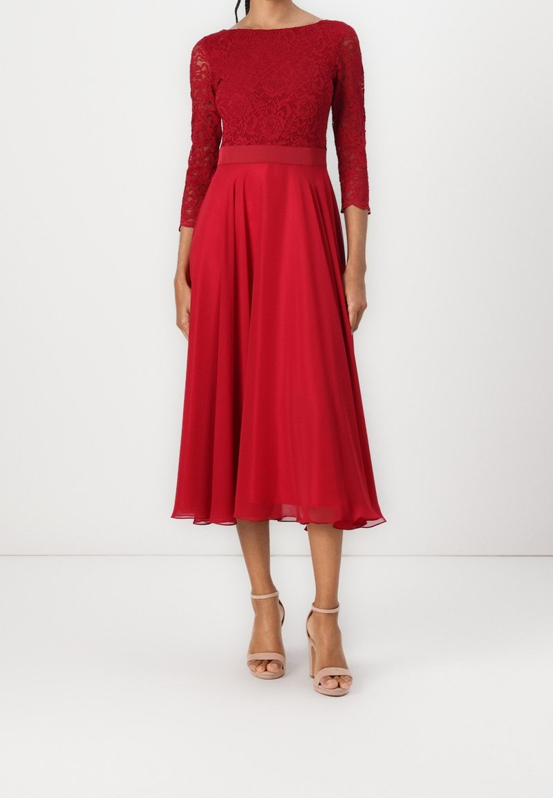 Robe rouge avec un corsage en dentelle, des manches trois-quarts et une jupe fluide en chiffon plissé. Associée à des talons nus à brides.