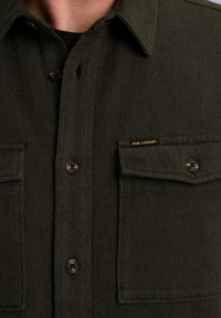 Camisa verde con patrón de espiga, dos bolsillos en el pecho, cierre de botones y logo de PME Legend en amarillo en el bolsillo izquierdo. Tejido texturizado.