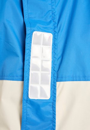 Modrá a béžová lehká bunda s vertikální průhlednou plastovou kapsou na boku, s texturovanou tkaninou a kontrastními švy.