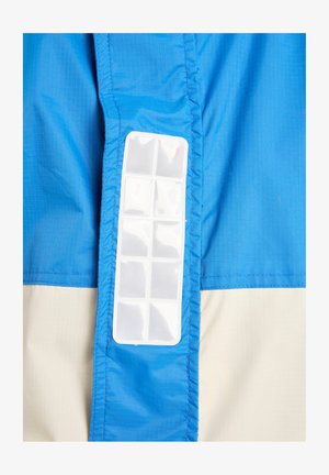 Svetlo modra in bež lahka jakna z navpičnim prozornim plastičnim žepom na strani, z teksturirano tkanino in kontrastnimi šivi.