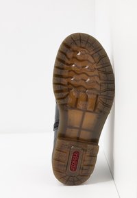 Sula av en svart sko med ett genomskinligt brunt gummiband och röd "rieker"-logotyp, placerad upprätt mot en vit hörnvägg.