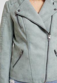 Veste de moto en suédine vert clair avec une fermeture éclair asymétrique, des poches latérales et des épaules matelassées.