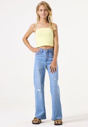 Lichtgele crop top met dunne bandjes, gecombineerd met lichtblauwe flared jeans met gescheurde details, en bruine sandalen met een accent.