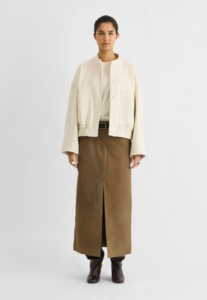 Le mannequin se tient devant un fond gris uni, vêtu d’une veste crème, d’une jupe marron longueur cheville et de bottes en cuir foncé.