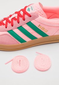 Tenisky Adidas Gazelle v růžové semišové úpravě, se zelenými pruhy a červenými tkaničkami. Součástí jsou dva sety kulatých růžových tkaniček.