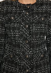 Schwarz-weiße kariert Tweedjacke mit tailliertem Design, ausgestattet mit metallischen Knöpfen und zwei Brusttaschen. Der strukturierte Stoff verleiht Tiefe.