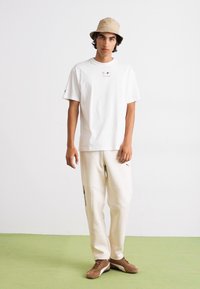 White short-sleeve t-shirt with a small logo, paired with cream-colored sweatpants featuring side zippers. Worn with a beige bucket hat and brown shoes.