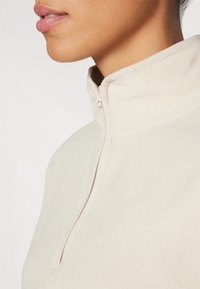 Pullover in felpa beige chiaro con colletto alto e dettagli con zip al collo, caratterizzato da una texture morbida e un design senza cuciture.