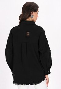Jachetă din denim neagră, supradimensionată, cu tiv destrămat și detalii rupte pe spate. Mâneci lungi cu manșete cu butoane.