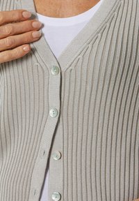 Grijze geribbelde cardigan met V-hals, voorzien van vijf knopen gemaakt van een lichtgekleurde schelp, gedragen over een wit shirt.