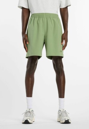 Personne portant un short vert clair à taille élastique, des chaussettes blanches et des chaussures de sport blanches, debout devant un fond uni.