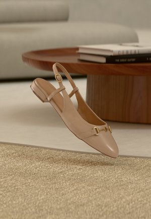 Beige læder slingback flad sko med gulddetalje på spænde, placeret i luften over et vævet tæppe, et træ sofabord og stablede bøger i baggrunden.