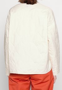 Krémová prošívaná bunda s texturovaným vzorem, kulatým výstřihem a dlouhými rukávy, doplněná oranžovými kalhotami.