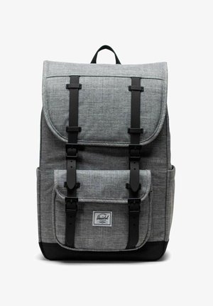 Sac à dos en tissu gris avec des sangles noires, poche frontale et poignée supérieure, présentant une étiquette blanche avec le nom de la marque sur la poche avant.