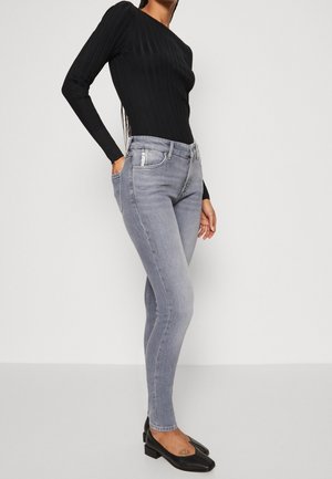 Frau trägt schwarzen, langärmligen, gerippten Pullover, hellgraue Skinny-Jeans und schwarze Schuhe mit niedrigem Absatz, steht mit einer Hand in der Tasche.