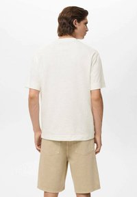 En mand står med ryggen til kameraet, iført en hvid kortærmet skjorte og beige shorts foran en ensfarvet baggrund.
