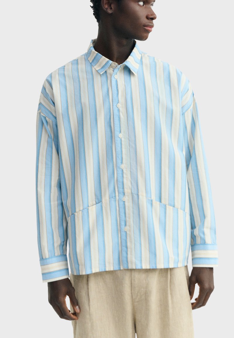 Junger Mann trägt ein locker sitzendes, langärmliges Hemd mit Knöpfen und vertikalen blauen, weißen und beigen Streifen, kombiniert mit hellfarbenen Hosen.