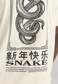 Kremo spalvos marškinėliai su juodu gyvatės grafiku, kuriame vaizduojamos detaliai matomos svarstyklės. Tekstas apima žodį "SNACK" ir informaciją apie Kinų zodiaką.