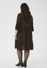 Rochie marron în etaje, cu decolteu rotund și mâneci evazate până la trei sferturi, având dungi subtile strălucitoare și o siluetă fluidă.