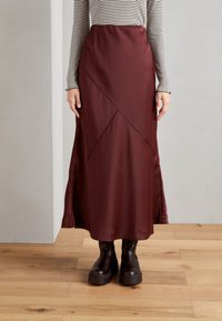 Fustă midi din satin bordeaux, cu cusături diagonale și o siluetă fluidă, asortată cu bocanci negri masivi, pe un podea din lemn.