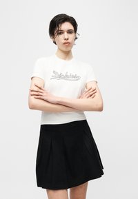 Γυναίκα με κοντά υγρά μαλλιά, σταυρωμένα χέρια, φορώντας λευκό κοντομάνικο πουκάμισο "Dickies" και μαύρη πλισέ φούστα μπροστά σε λευκό φόντο.