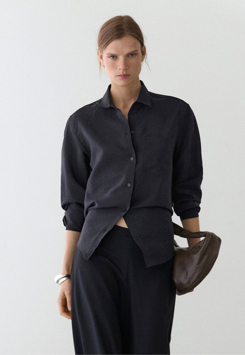 Jeune femme aux cheveux châtain clair porte une chemise noire ample à boutons et un pantalon noir, tenant un sac à main en cuir marron dans sa main gauche.