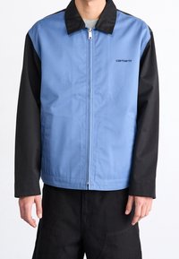 Blå og sort jakke med fuld lynlås, med en opretstående krave og sidelommer lavet af et glat stof. Logo på brystet.